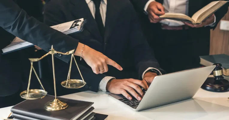 Lawyers collaborating at a desk, with one pointing at a laptop screen beside legal documents and scales