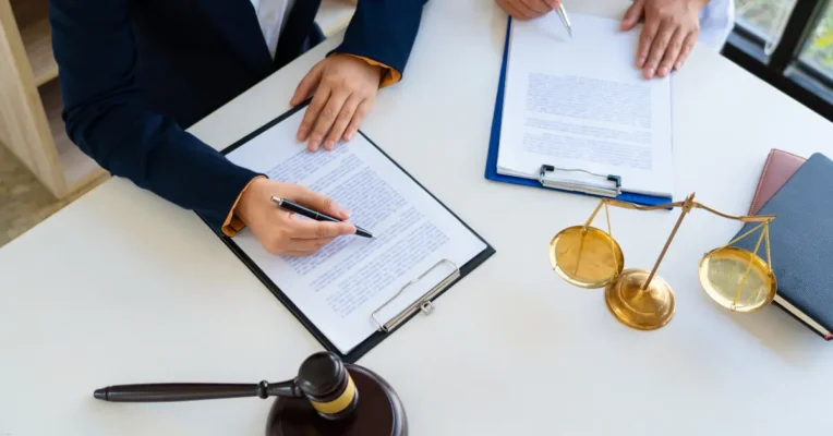 trust lawyer signing a document beside scales of justice and a gavel on a white desk